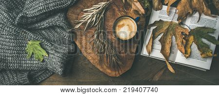 Autumn morning coffee concept. Flat-lay of knitted woolen jamper, wooden tray, mug of coffee, yellow fallen leaves and book over dark rustic wooden table background, top view