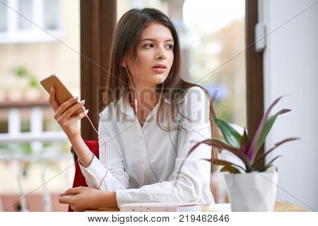 Beautiful young woman using her smart phone wearing ear phones looking away thoughtfully technology device gadget communication conncetivity mobility carrier concept.