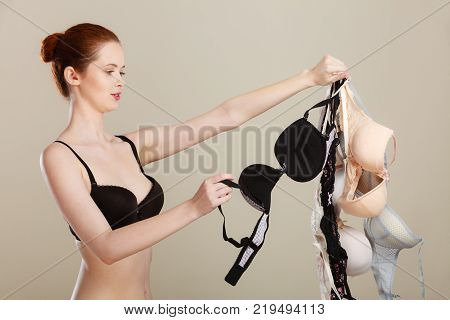 Bosom concept. Slim attractive red hair woman wearing black underwear holding many bras in hand choosing witch bra to wear