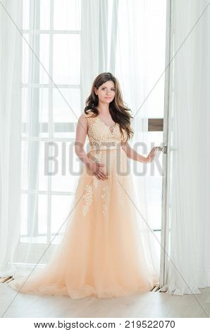 pregnant brunette woman in a ball cream gown staya near a balcony door
