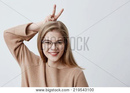 Portrait of cute adorable female model in spectacles looks joyfully at camera, gestures with fingers, has happy expression, poses against gray background with copy space. Pretty blonde girl having fun