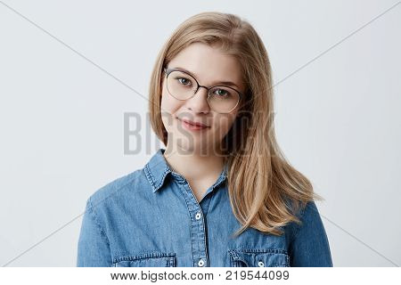 Portrait of young tender blonde teenage girl with healthy skin wearing denim shirt and spectackles looking at camera with pleased or pensive expression. Caucasian woman model with blonde hair posing indoors