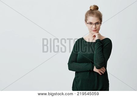 Pretty young female looks with appeal at camera having her blonde hair tied in knot wearing big glasses and green loose sweater holding hand under her chin, building plans, thinking about something