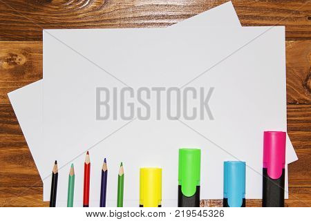 A white sheet of paper lay on a wooden table, near , pencils, markers, pens . The view from the top