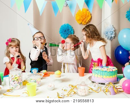 Group of smiling children playing on the birthday party in decorated room. Happy kids blowing in pipes on birthday party