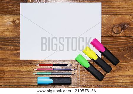 A white sheet of paper lay on a wooden table, near , pencils, markers, pens . The view from the top