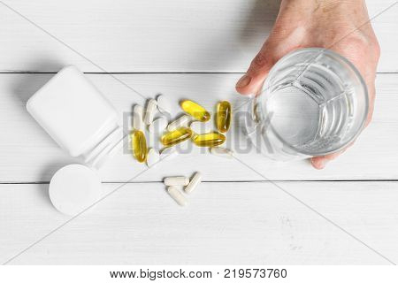 Woman hands takes yellow capsules of omega 3 white pills of calcium glucosamine supplements glass of water and plastic bottle on wooden planks table.