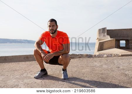 Portrait of calm young Latin American athlete wearing orange t-shirt sitting on hunkers after training on beach. Fitness and wellbeing concept