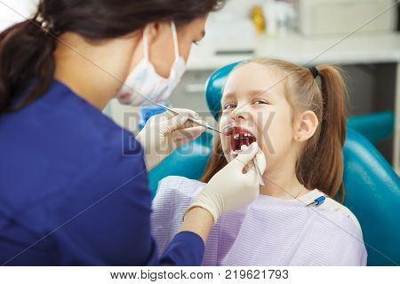 Child lies in comfortable dentis chair and goes through procedure without fear while female doctor in mask and rubber gloves fix girls teeth in modern medical office.
