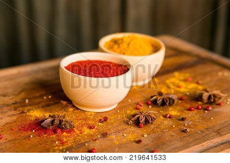 Close up two bowls filled with red paprika and yellow turmeric powder on dark wooden table covered with anise