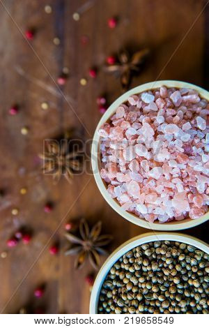 Pink Himalayan salt and coriander seeds in white bowls on wooden surface covered with anise and red peppercorn