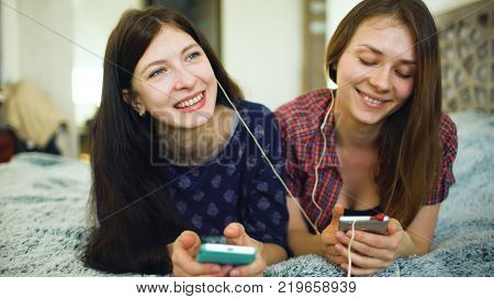 Two yuong woman friends listening to music with headphones connected to smarhphone lie on bed at home