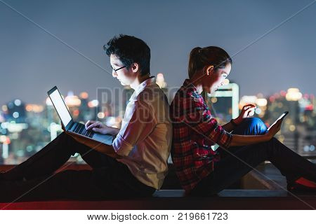 Young Asian couple using laptop and smartphone together, lean on each other back to back. Rooftop night scene with city view background. Modern technology lifestyle or internet gadget devices concept