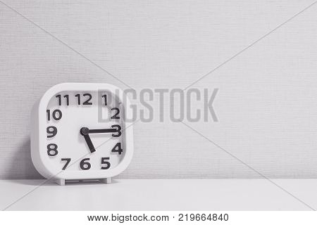 Closeup white clock for decorate show a quarter past five o'clock or 5:15 p.m. on white wood desk and cream wallpaper textured background in black and white tone with copy space