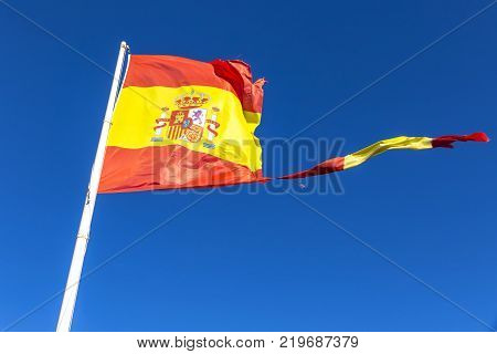 Close-up torn Spanish flag waving on the wind on the mast over clear blue sky background