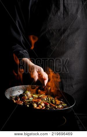 Chef cooking vegetables on a pan with fire. Black background for copy text. Vertical photo for banner design.