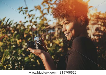 Squinting cute young African American female tourist with old retro camera during photographing; admirable black girl with vintage photo camera tiking pictures in public park on a sunny evening