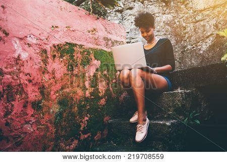 Young lovely Brazilian female student is sitting on a stony staircase near reddish mossy wall with laptop; cute black undergraduate girl with net-book on a step of stone stairs is making her homework