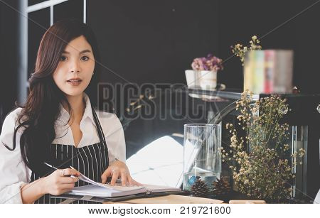 small business owner with notebook at counter in coffee shop. asian female barista wearing apron at bar in cafe. food service restaurant entrepreneur concept.
