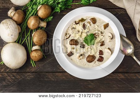 An overhead photo of a plate of a mushroom cream soup with champignons, cremini, fresh parsley, on a dark rustic background texture with a place for text