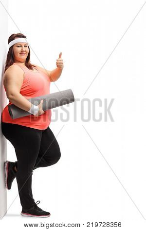 Full length portrait of an overweight woman with an exercise mat making a thumb up sign and leaning against a wall isolated on white background
