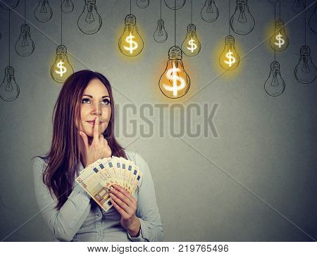 Portrait thinking young woman holding money looking up at dollar idea light bulbs above head