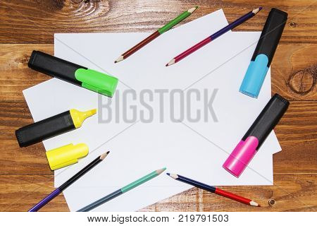 A white sheet of paper lay on a wooden table, near , pencils, markers, pens . The view from the top
