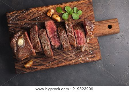 Closeup ready to eat steak new York beef breeds of black Angus with herbs, garlic and butter on a wooden Board. The finished dish for dinner on a dark stone background. Top view.