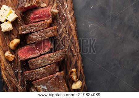 Closeup ready to eat steak new York beef breeds of black Angus with herbs, garlic and butter on a wooden Board. The finished dish for dinner on a dark stone background. Top view with copy space.