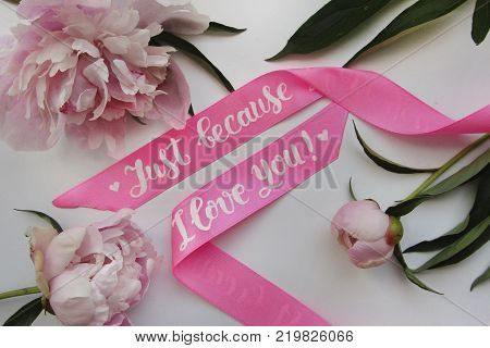 Flat lay composition with satin pink ribbon with modern calligraphy inscription `just because I love you` and peonies on white background for postcard, poster