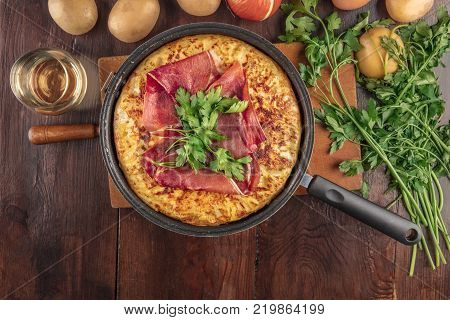 An overhead photo of a Spanish tortilla in a traditional tortillera, with jamon and parsley, on dark rustic textures with wine, ingredients, and a place for text