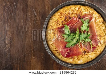 An overhead closeup photo of a Spanish tortilla in a traditional tortillera, with jamon and parsley, on dark rustic textures with a place for text