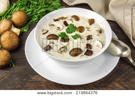 A closeup photo of a plate of a mushroom cream soup with champignons, cremini, fresh parsley, and pepper, on a dark rustic background