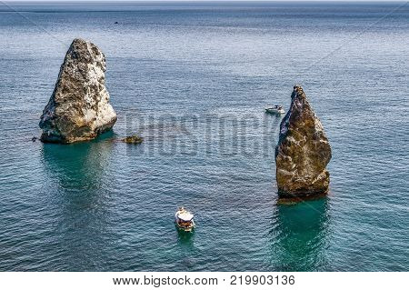Two sharp rocks in the crystal clear emerald sea with white pleasure craft. The view from the top.