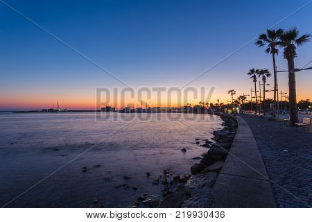 View over the seafront of the city Paphos in Cyrpus. Palm silhouettes, boats in the water and golden sunset over the sea with dramatic cloudy sky.
