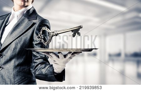 Cropped image of waitress's hand in white glove presenting big key on metal tray with office view on background.