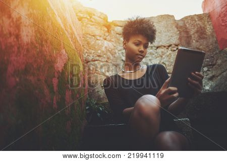 Thoughtful young curly Brazilian undergraduate female is sitting on stone staircase outside of her college and surfing internet via digital pad, with copy space zone for advert, message or your logo