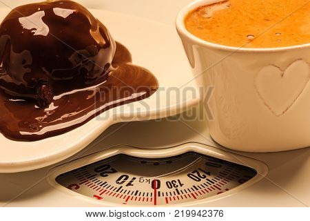Cup of coffee and chocolate cake on heart shaped plate saucer on weighing scale. Unhealthy nutrition. Fat diet concept.