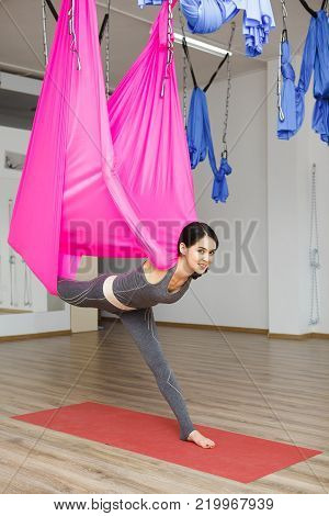 Girl stretching leg with help of hammock. Aerial exercise or antigravity yoga indoors, woman does aero poses with head leaned to back
