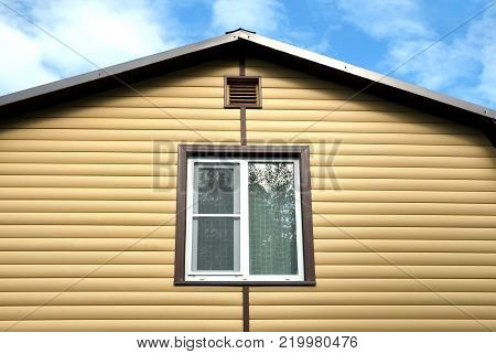 Upper floor with plastic window  in country house wall covered with yellow siding and brown metal roof on blue sky with clouds on sunny day front view closeup