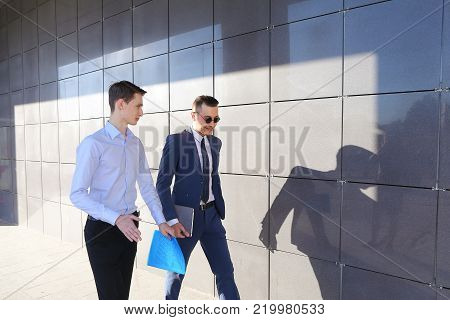 Two attractive confident young businessman male guys go and discuss important issues, hurry to a meeting, make decisions, solve problems, share business ideas, smiling, holding colored folders in hands, go along gray walls of modern business center outdoo