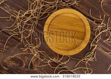 Lot Of Pieces Of Thin Raw Pasta Noodles With Empty Bamboo Plate Flatlay On Brown Wood
