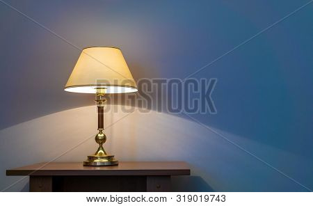 Classic Table Lamp With Turned On Bulb Under Lampshade Is Against Wall With Blue Wallpaper. Copy Spa