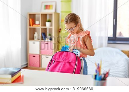 children, education and learning concept - happy student girl in glasses packing school bag at home