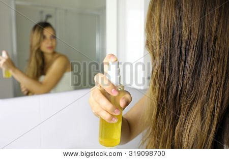 Woman Applying Oil Mask Spray On Hair In Front Of A Mirror. Hair Care Concept. Focus On Hair.