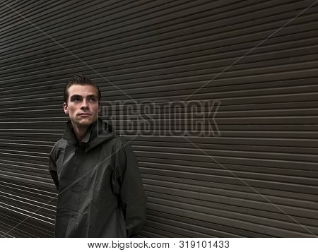 Handsome Young Man In A Raincoat Stands In Front Of A Wall, Garage Doors. Cope Space, Place For Text