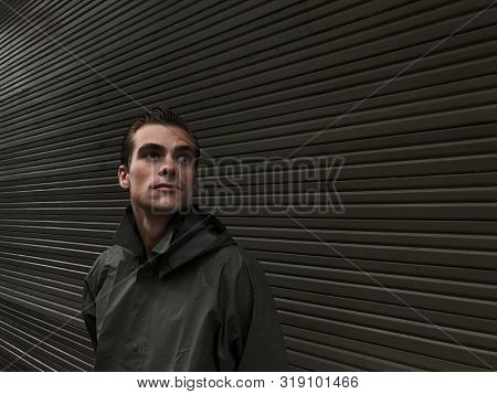Handsome Young Man In A Raincoat Stands In Front Of A Wall, Garage Doors. Cope Space, Place For Text