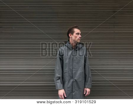 Handsome Young Man In A Raincoat Stands In Front Of A Wall, Garage Doors. Cope Space, Place For Text