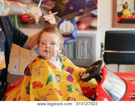First Baby Haircut. Cute Blonde Toddler Is Happy To Be On A Haircut With A Professional Baby Hairdre