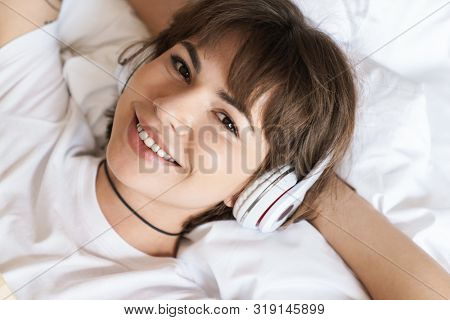 Photo of a happy optimistic positive pleased young beautiful woman lies indoors at home on bed listening music with headphones looking camera.